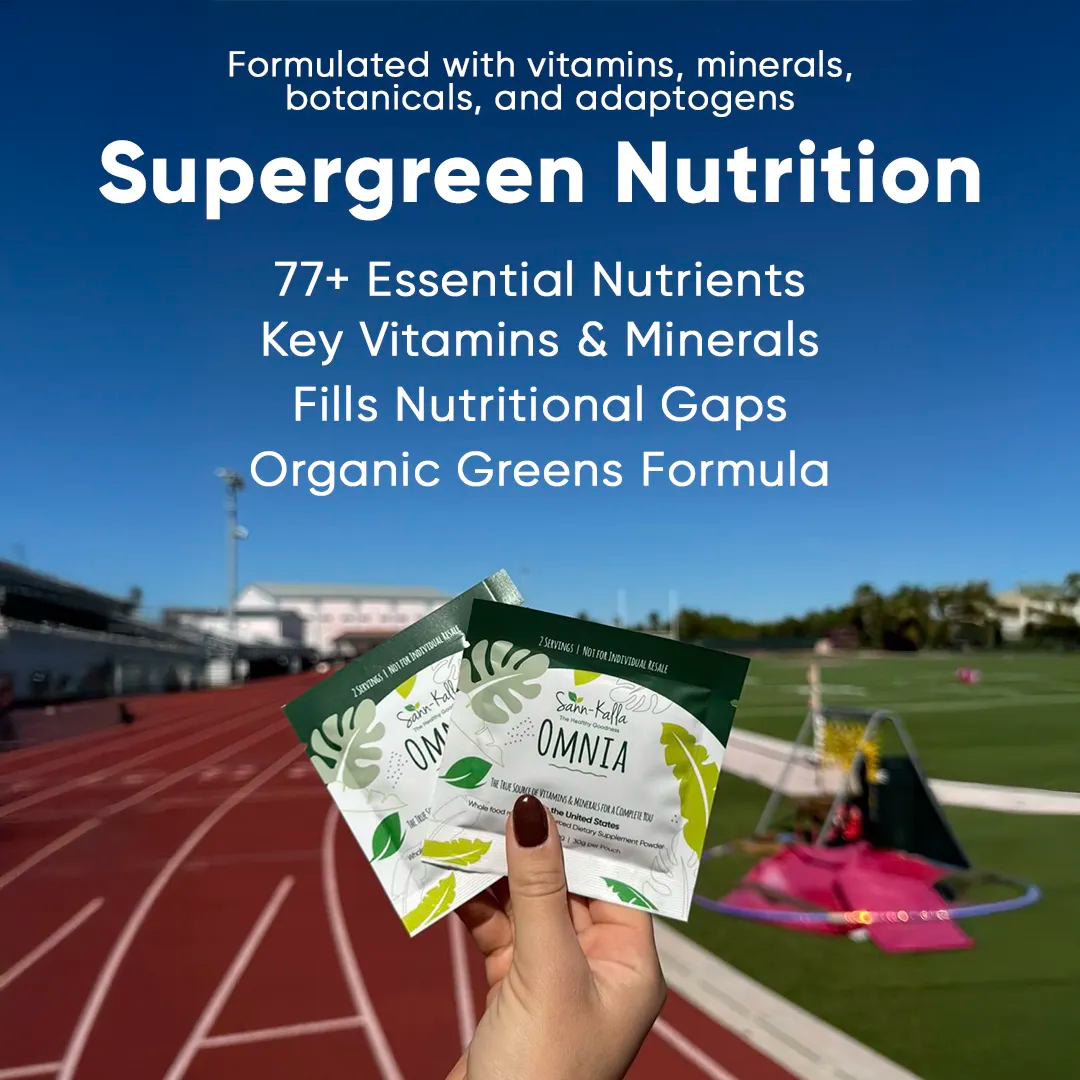 Sann-Källa Omnia Supergreen Nutrition sample packs held outdoors at a track field, showcasing a supergreens drink mix formulated with 77+ essential vitamins, minerals, botanicals, and adaptogens to help fill nutritional gaps and support everyday wellness.